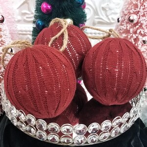 May include: Three red knitted Christmas ornaments with a cable knit pattern. The ornaments are displayed on a silver tray with rhinestones.