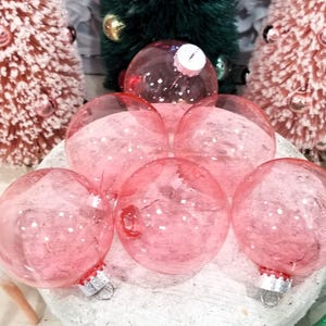 May include: A collection of pink Christmas ornaments with silver caps and glitter inside. The ornaments are arranged on a white surface, with pink and green decorative trees in the background. The ornaments are round and transparent.
