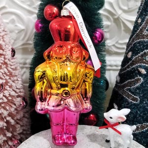 May include: A colorful Christmas ornament shaped like a toy soldier, with a gradient of red, gold, and pink. The ornament has a metallic finish and a white ribbon that says "Love, Dominic". A small white deer figurine with a red bow is also present.