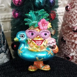 May include: A whimsical pineapple ornament wearing pink glitter sunglasses and a flower crown, floating in a blue duck-shaped inner tube. The ornament is holding a small ice cream cone and wearing sandals. Christmas tree ornaments are in the background.