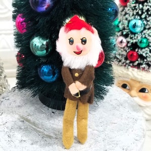 May include: A vintage felt gnome ornament with a red hat, white beard, brown jacket, and yellow pants. The gnome is standing in front of a green artificial Christmas tree.