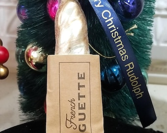 May include: A gold glass ornament shaped like a baguette with a brown paper bag label that reads "French Baguette". The ornament is hanging from a blue ribbon with the text "Merry Christmas Rudolph".
