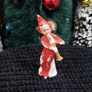 May include: A small ceramic figurine of an elf wearing a red pointed hat and a red suit with white accents. The elf is depicted in a standing position, holding a gold-colored trumpet to its mouth. The paint on the red suit shows signs of wear.