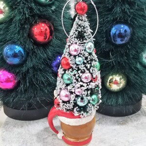 May include: A Christmas tree ornament shaped like a tree atop a mug. The tree is decorated with colorful ornaments and dusted with snow. The mug has a red handle and trim, with a white band around the middle. Other Christmas trees with ornaments are in the background.