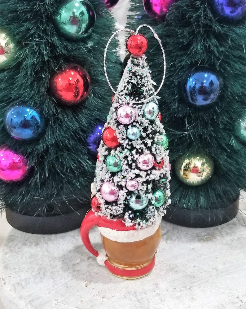 May include: A Christmas tree ornament shaped like a tree atop a mug. The tree is decorated with colorful ornaments and dusted with snow. The mug has a red handle and trim, with a white band around the middle. Other Christmas trees with ornaments are in the background.