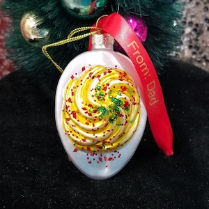 May include: A white, egg-shaped Christmas ornament with a yellow and gold swirl design, red and green glitter accents, and a red ribbon that says "From: Dad". The ornament hangs from a gold string.