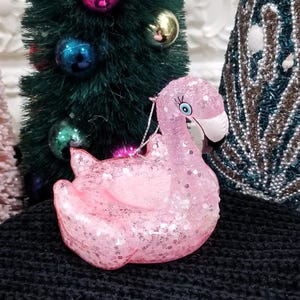 May include: A pink flamingo Christmas ornament covered in glitter. The ornament has a white beak, blue eye, and a silver string for hanging. The ornament is sitting on a black surface, with a Christmas tree in the background.