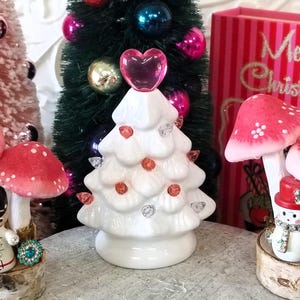 May include: A white ceramic Christmas tree with a pink heart topper and orange and clear gem accents. The tree is surrounded by other Christmas decorations, including a snowman figurine and mushroom ornaments.