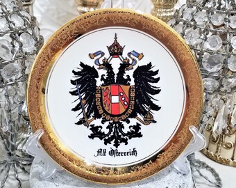 May include: A white porcelain plate with a gold rim and a detailed floral pattern. The center features a black and gold coat of arms with a red shield and the text "Alt Osterreich".