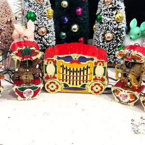 May include: A collection of six miniature carousel animal ornaments and a central red and gold circus wagon ornament. Each carousel piece features a different animal, including an elephant, tiger, and zebra, with gold, red, and green accents.