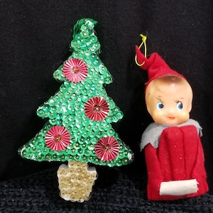 May include: A Christmas tree ornament covered in green sequins with red pleated circular accents and a gold sequined base, positioned next to a small elf figurine wearing a red felt hat and suit with a grey collar and a white trim detail.