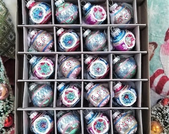 Puede incluir: Un juego de 24 adornos navideños vintage de vidrio en una caja de cartón. Los adornos son plateados con acentos azules, verdes y rosados. Cada adorno tiene un diseño diferente.