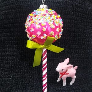May include: A pink Christmas ornament shaped like a lollipop, covered in colorful beads and topped with a green bow. A small pink deer figurine with a red bow is next to the ornament.