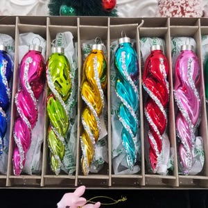 May include: A set of eight colorful, vintage Christmas tree ornaments in a cardboard box. Each ornament is a twisted, teardrop shape with a silver glitter accent. Colors include blue, pink, green, yellow, teal, red, and purple.