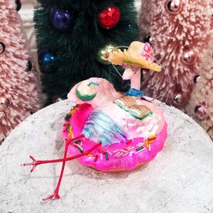 May include: A whimsical pink flamingo Christmas ornament. The flamingo is perched on a pink donut with colorful sprinkles. It wears a straw hat and has a book and drink. The ornament is set against a backdrop of miniature Christmas trees.