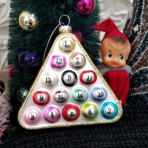 May include: A Christmas ornament shaped like a pool rack, featuring numbered billiard balls in various colors. The ornament has a gold trim and is hanging near a small doll wearing a red outfit and hat.