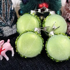 May include: Four round, light green Christmas ornaments with silver clips and green beaded trim. A small pink deer figurine with a red bow is also present. The ornaments have a glittery texture, suggesting a festive holiday theme.