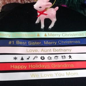 May include: A pink reindeer ornament with a red bow sits atop several ribbons. The ribbons are in various colors, including green, blue, pink, white, and red, and feature holiday greetings and messages.