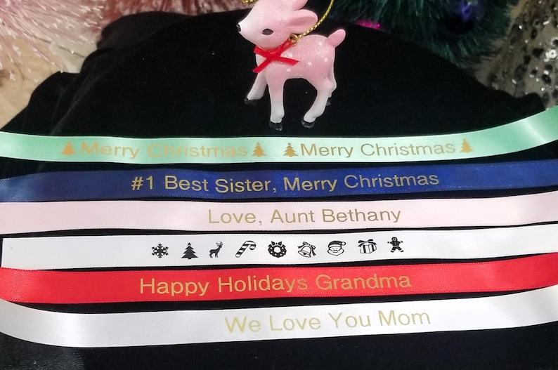 May include: A pink reindeer ornament with a red bow sits atop several ribbons. The ribbons are in various colors, including green, blue, pink, white, and red, and feature holiday greetings and messages.