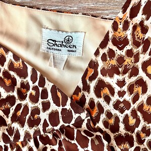 May include: A vintage dress with a brown and orange leopard print pattern. The dress has a V-neck design and a tag that reads "Shaheen California Hawaii". The fabric appears to be a lightweight material.