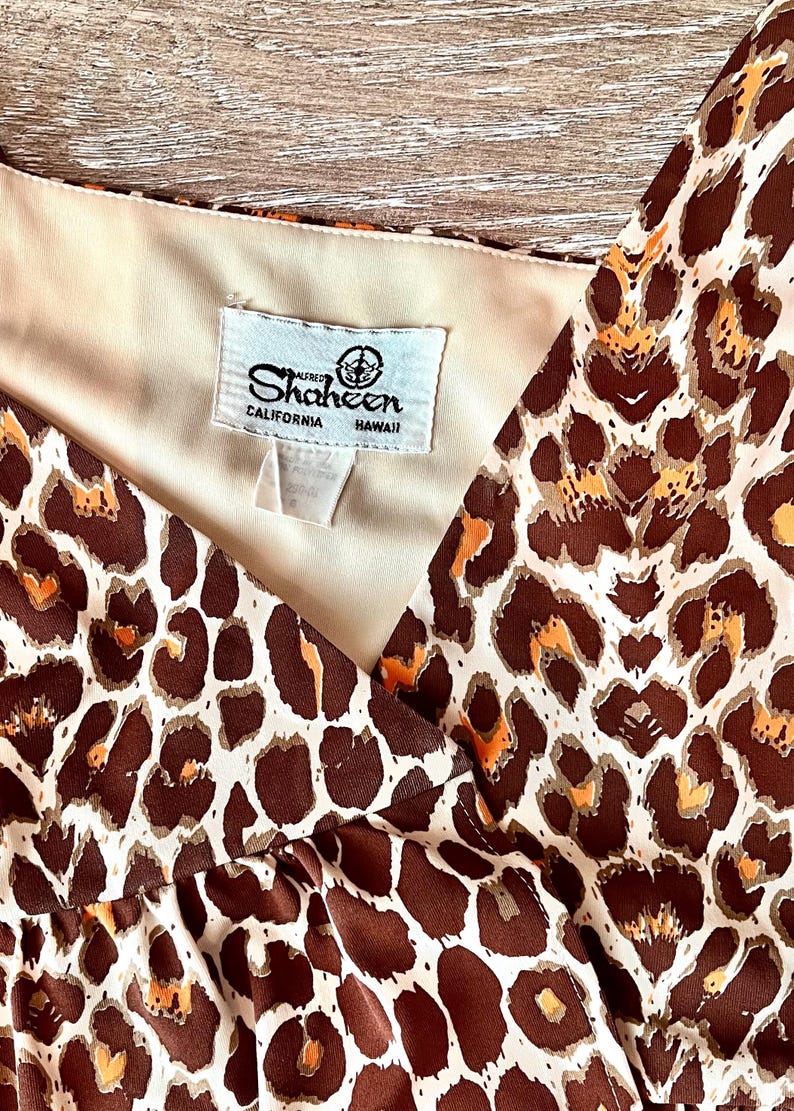 May include: A vintage dress with a brown and orange leopard print pattern. The dress has a V-neck design and a tag that reads "Shaheen California Hawaii". The fabric appears to be a lightweight material.