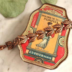 May include: A copper-toned bracelet with leaf-shaped charms rests on a vintage talc tin. The tin features a red and yellow label with the text "POUDRE DE TALC POMPEIA" and an illustration of a woman. The bracelet is connected with a clasp.