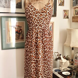 May include: A vintage-style dress with a leopard print pattern. The dress features a V-neckline, spaghetti straps, and a fitted waist. The brown and tan animal print is displayed on a dress form in a room with framed artwork.