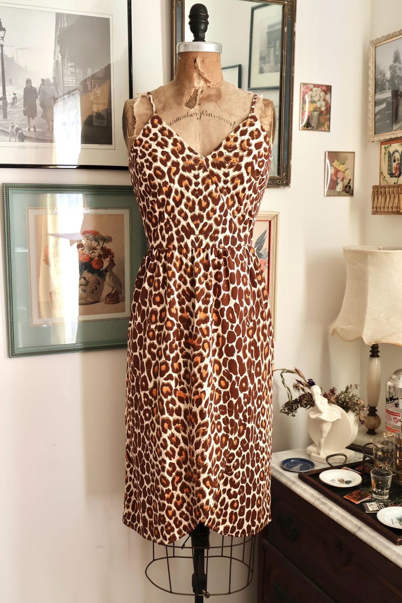 May include: A vintage-style dress with a leopard print pattern. The dress features a V-neckline, spaghetti straps, and a fitted waist. The brown and tan animal print is displayed on a dress form in a room with framed artwork.