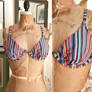 May include: A vintage-style, multi-colored striped bikini top. The top features vertical stripes in various colors, including red, blue, yellow, and black. The bra is displayed on a vintage mannequin. The straps are thin and adjustable.
