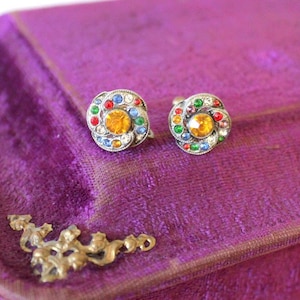 Puede incluir: Un par de pendientes de clip de tono plateado con un diseño floral. Los pendientes están adornados con brillantes de colores en tonos rojo, naranja, amarillo, verde, azul y blanco.