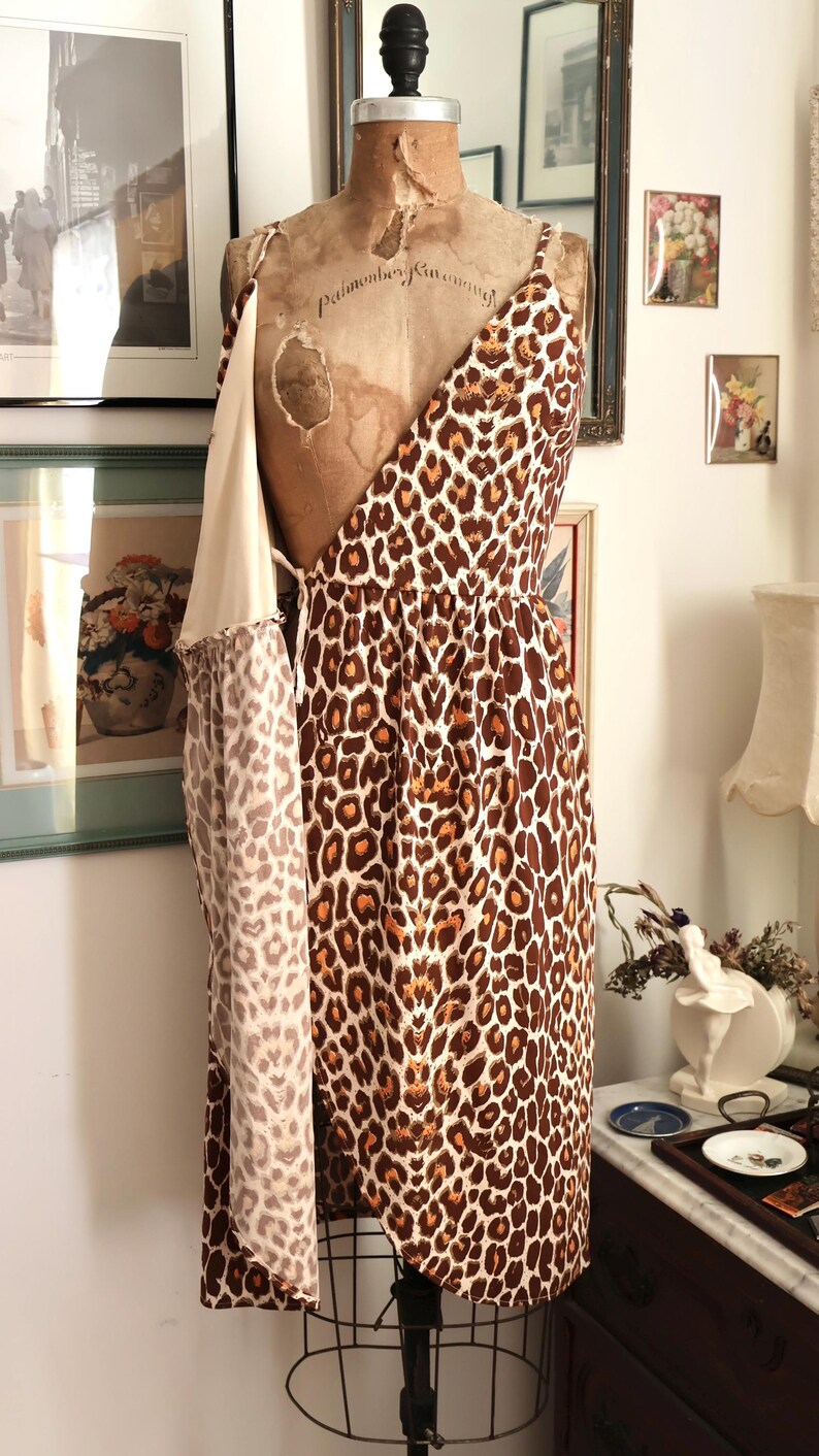 May include: A vintage leopard print dress on a mannequin. The dress features a brown and cream animal print pattern. The dress has spaghetti straps and a draped design. The mannequin is a vintage style with a cream-colored torso.