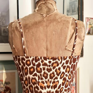 May include: A brown and white leopard print sundress on a vintage dress form. The dress has thin straps and a fitted bodice. The dress form is a light brown color and has a worn appearance. The background includes framed artwork and a mirror.