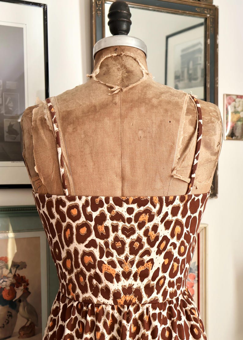 May include: A brown and white leopard print sundress on a vintage dress form. The dress has thin straps and a fitted bodice. The dress form is a light brown color and has a worn appearance. The background includes framed artwork and a mirror.