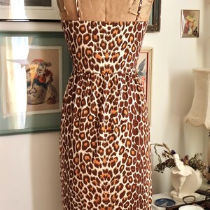 May include: A vintage-style dress on a mannequin, featuring a brown and white leopard print. The dress has thin straps and a fitted bodice that flares into a knee-length skirt. The dress is displayed in a room with framed artwork.
