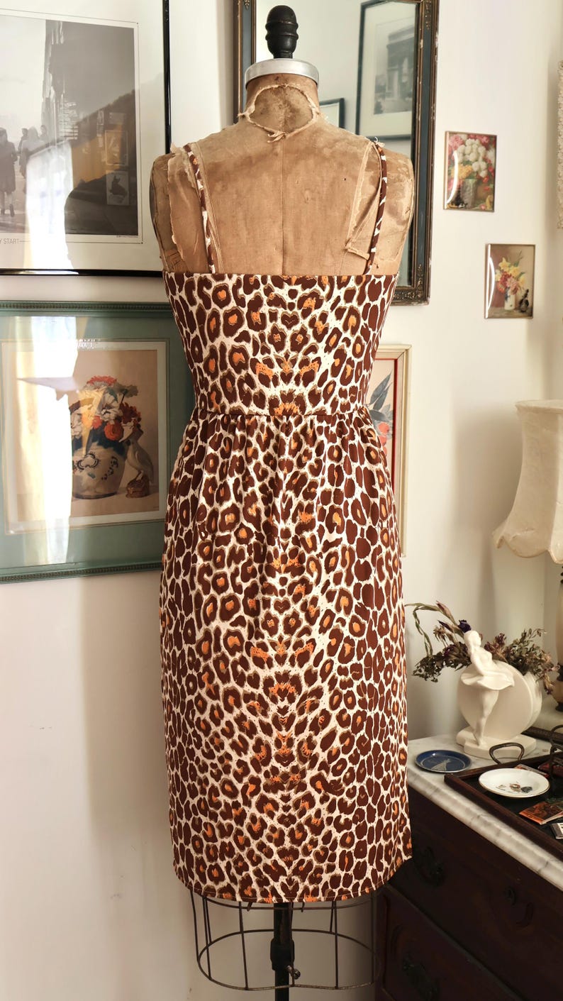 May include: A vintage-style dress on a mannequin, featuring a brown and white leopard print. The dress has thin straps and a fitted bodice that flares into a knee-length skirt. The dress is displayed in a room with framed artwork.