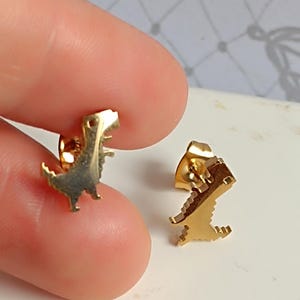 Puede incluir: Un par de pendientes de botón dorados con un diseño de dinosaurio pixelado.