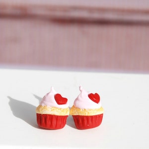 Puede incluir: Dos cupcakes en miniatura con bases rojas, capas de bizcocho amarillas y glaseado rosa rematado con un corazón rojo. Los cupcakes están sobre una superficie blanca con un fondo rosa suave, lo que sugiere un tema de San Valentín.