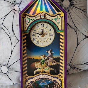 May include: A rectangular wall clock with a purple frame and a pointed top. The clock face features Roman numerals. Below, a scene depicts two figures riding horses. The text reads "CAROUSEL" and "The clowns began to sing to cheer the way."
