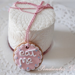 May include: A white cake with an intricate embossed design, paired with a pink-frosted cookie that reads "eat me" in white script. A pink beaded chain is draped over the cake and cookie, creating a whimsical, dessert-themed presentation.