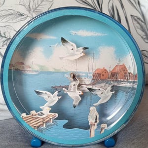 Puede incluir: Una pieza decorativa redonda, con borde azul, que presenta una escena costera con gaviotas en vuelo. La obra de arte representa un puerto con barcos, edificios y un muelle. La pieza está sostenida por dos pequeños pies azules.