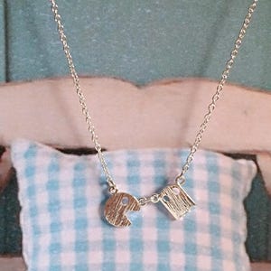 May include: A silver necklace with two silver charms shaped like Pac-Man ghosts. The necklace is on a white and blue checkered pillow.