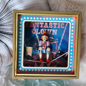 Puede incluir: Una caja de sombra cuadrada con marco dorado que presenta una figura de payaso en miniatura equilibrándose en una cuerda floja. El fondo muestra una escena de circo con el texto "FANTASTIC CLOWN". El payaso viste un traje rosa y azul.