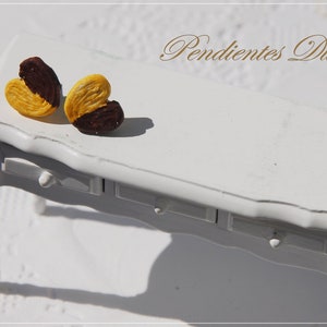 May include: Two heart-shaped earrings made of polymer clay. The earrings are painted to look like a palmier cookie, with a brown chocolate center and a yellow pastry exterior. The earrings are displayed on a white table with the text "Pendientes Dulces" in the background.