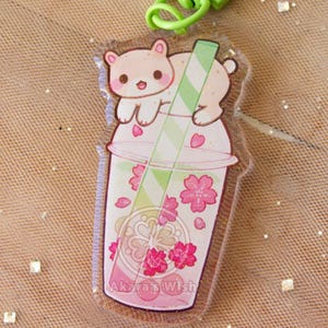 May include: A clear acrylic keychain featuring a pink drink with cherry blossoms and a cute bear illustration. The keychain has a green clasp and a green and white striped straw. The text "Akara's Wish" is visible.