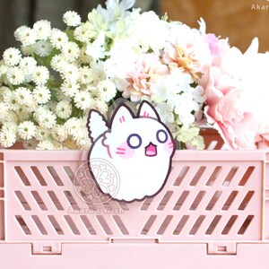 May include: A white, cartoon cat with pink cheeks and a surprised expression is printed on a clear acrylic charm. The charm is sitting in a pink plastic crate with white flowers.