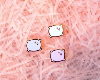 Puede incluir: Tres pines de esmalte rosa, blanco y morado con forma de almohadas con ojos brillantes. Los pines están sobre un fondo de papel de seda rosa arrugado. Akara's Wish está impreso en la esquina superior derecha de la imagen.