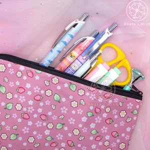 Può includere: Un astuccio per matite rosa con cerniera e un motivo floreale bianco e verde con fragole. L'astuccio è pieno di penne, matite e un paio di forbici.