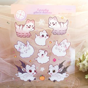 May include: A sheet of stickers featuring cartoon kittens in various Halloween costumes. The stickers include ghost kittens, kittens with bat wings, and kittens with multiple eyes. The stickers are in shades of white, pink, and gray, with small details like candy corn and spiderwebs.