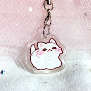 Könnte beinhalten: Ein transparenter Schlüsselanhänger mit einer weißen Katzenillustration. Die Katze hat rosa Wangen und ist von goldenem Glitzer umgeben.