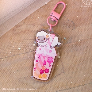 Op de afbeelding: Een roze en witte bubble tea-hanger met een klein schaapje dat er bovenop zit. De hanger is gemaakt van helder acryl en heeft een roze en wit gestreepte rietje. De hanger is bevestigd aan een roze metalen sleutelhanger.
